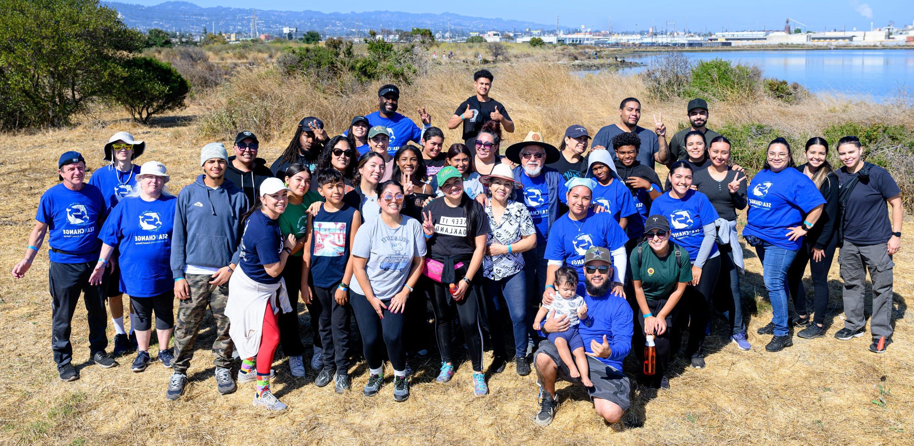 A large group of volunteers, all friends and family of one employee, smile for the camera on the shoreline of the Oakland Estuary.